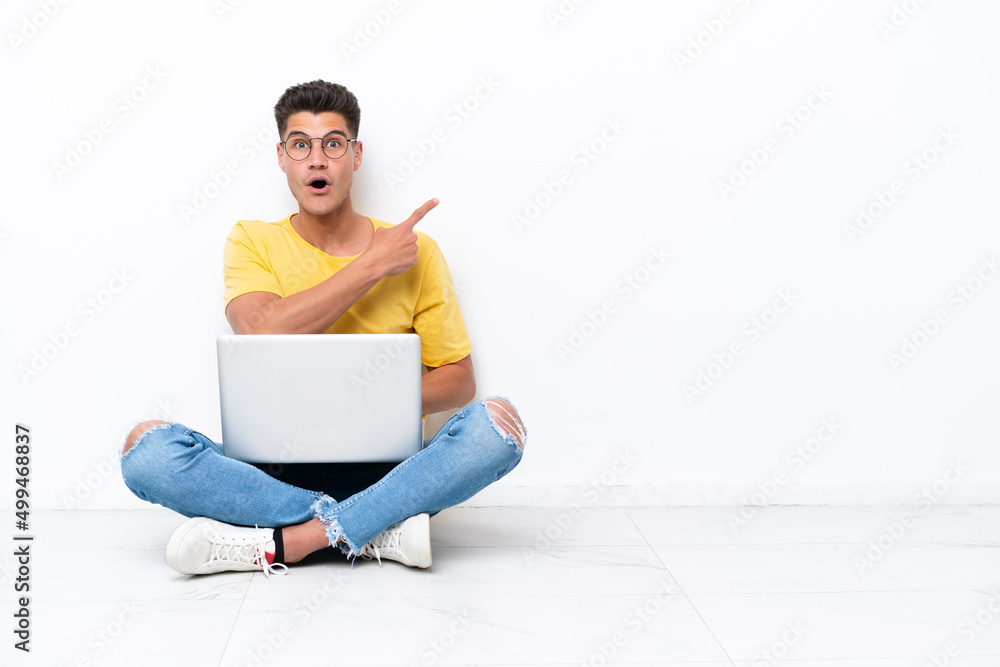 Young man sitting on the floor isolated on white background surprised and pointing side Young man sitting on the floor isolated on white background surprised and pointing side
