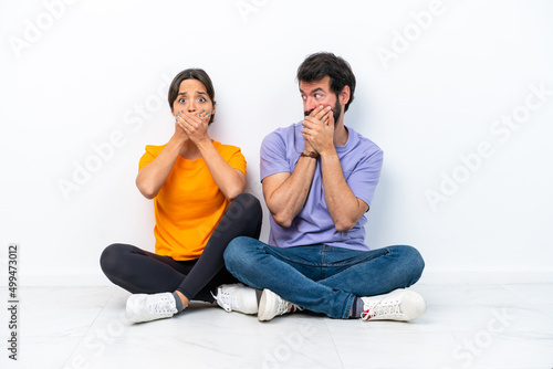 Young caucasian couple sitting on the floor isolated on white background covering mouth with hands