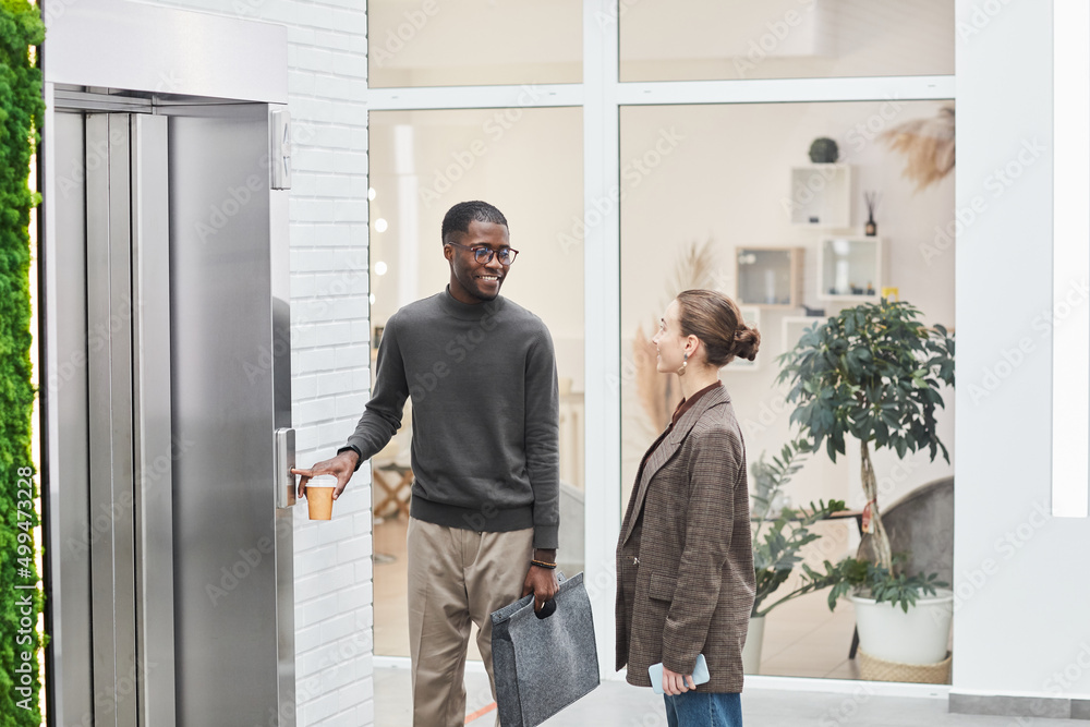 Side view portrait of two young coworkers standing by elevator in modern office building and chatting Side view portrait of two young coworkers standing by elevator in modern office building and chatting