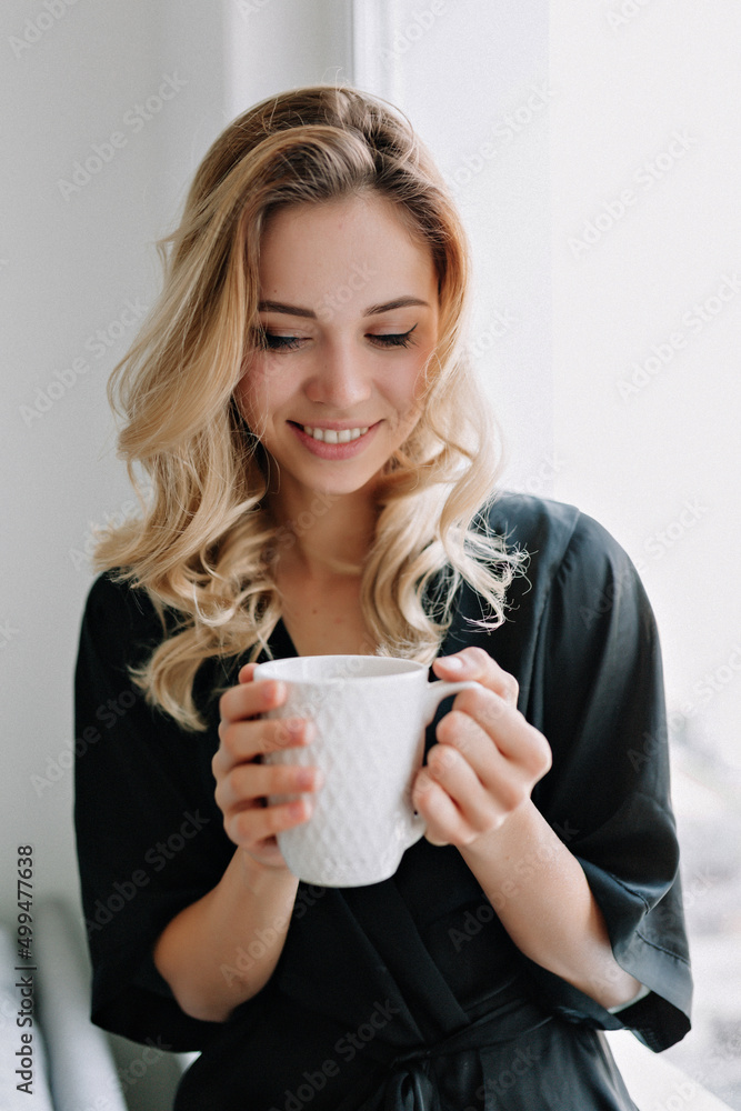 Relaxed adorable smiling girl is smiling and holding morning cup of coffee. Indoor shot of amazing lady sitting at home and resting near the window Relaxed adorable smiling girl is smiling and holding morning cup of coffee. Indoor shot of amazing lady sitting at home and resting near the window