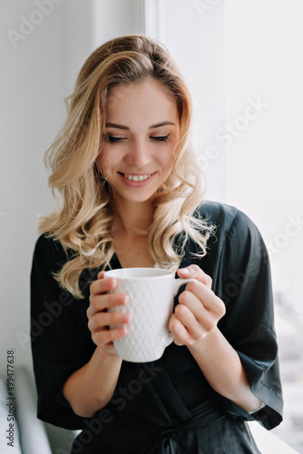 Relaxed adorable smiling girl is smiling and holding morning cup of coffee. Indoor shot of amazing lady sitting at home and resting near the window