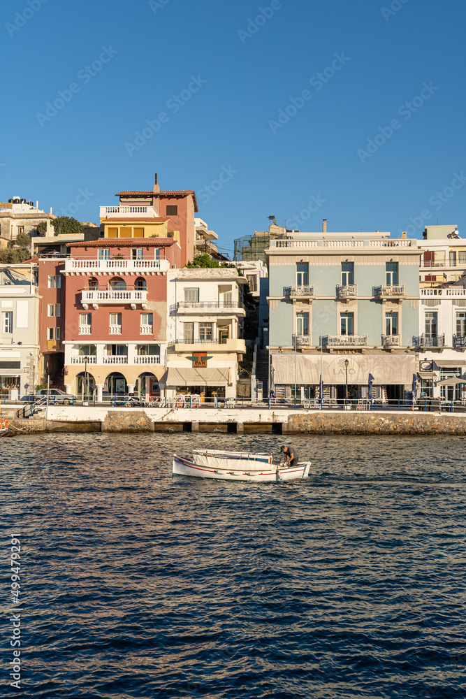 Ein kleines Boot fährt durch den Hafen von Agios Nikolaos auf Kreta, Griechenland, im Hintergrund farbenfrohe Häuser, direkt am Wasser des Hafen gebaut