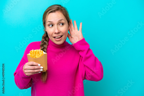 Young caucasian woman holding fried chips isolated on blue background listening to something by putting hand on the ear