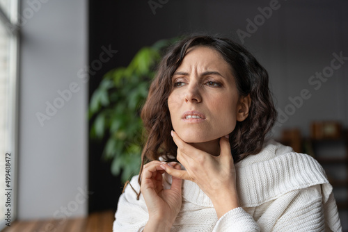 Young sick worried woman with sore throat at home touching neck, unhealthy female wrapped in plaid having swollen lymph nodes caused by bacterial infection, feeling pain or discomfort while swallowing photo
