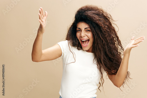 Happy joyful young Latin woman in casual gray sweatshirt doing winner gesture celebrating clenching fists say yes look to you isolated on beige color background studio portrait. Copy space for ad