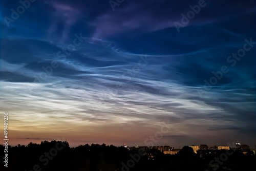 Noctilucent clouds in night sky. Rare atmospheric phenomenon over city. Shining clouds over silhouette of night town.