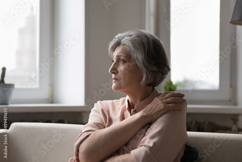 Free Download Older woman sit on couch in living room staring into distance feeling lonely, suffers from solitude. Sad mature 60s female reminisces past, looks unhappy, misses grown up children looks upset concept - GFXLoad