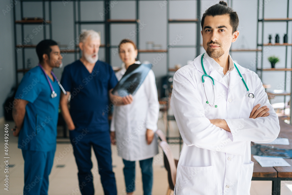 Portrait of handsome young male doctor in white uniform standing in medical meeting office, looking at camera. Multiethnic team of physician working in background, discussing MRI scan of patient. Portrait of handsome young male doctor in white uniform standing in medical meeting office, looking at camera. Multiethnic team of physician working in background, discussing MRI scan of patient.