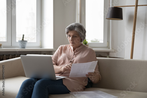 Older grey-haired woman sits on sofa with laptop pay bills through online e-bank app looks serious, do paperwork, manage personal finances, analyze papers, makes payments, check utility rates concept