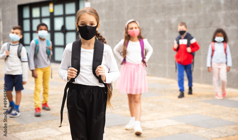 Obraz premium Portrait of tween schoolgirl in protective face mask with rucksack on her way to college on warm fall day. Back to school after lockdown concept.