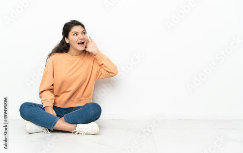Teenager Russian girl sitting on the floor listening to something by putting hand on the ear