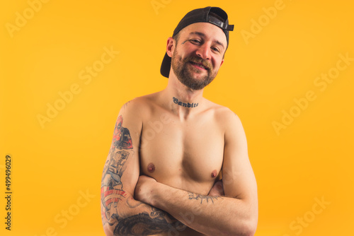 Obstreperous gangster-like bad bare-chested caucasian middle-aged man in a black cap with visor on the back of his head looking at camera over yellow background. High quality photo