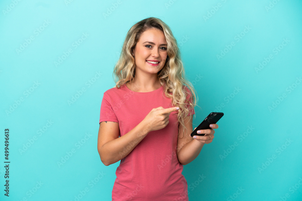 Young Brazilian woman isolated on blue background using mobile phone and pointing it Young Brazilian woman isolated on blue background using mobile phone and pointing it