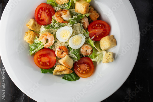 Caesar salad with grilled shrimps, tomato, boiled egg, lettuce, croutons and green sauce. Top view close-up on a brown round plate with fresh vegetables and cutlery by side on a dark background.