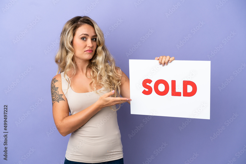 Young Brazilian woman isolated on purple background holding a placard with text SOLD and pointing it Young Brazilian woman isolated on purple background holding a placard with text SOLD and pointing it