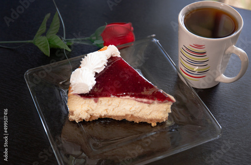 A slice of cheesecake in a glass plate with a coffe cup and a rose