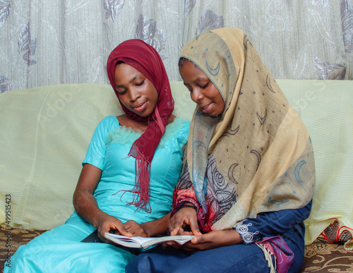 Two happy African Nigerian female muslim sisters or friends with Hijab scarf, sitting together while studying with a book placed on their laps