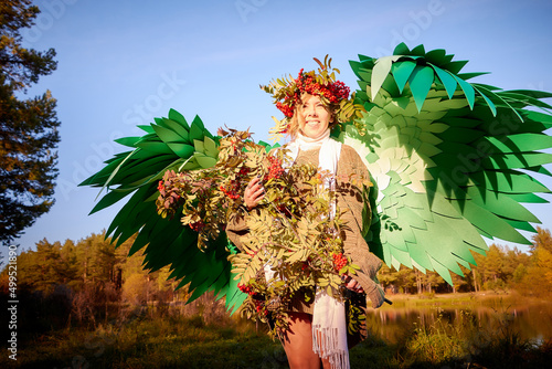 Cute girl in sweater, white scarf and green wings with bouquet of rowan branches with red berries and wreath in nature on sunny autumn. Model poses in green and yellow landscape as elf, dryad or angel photo
