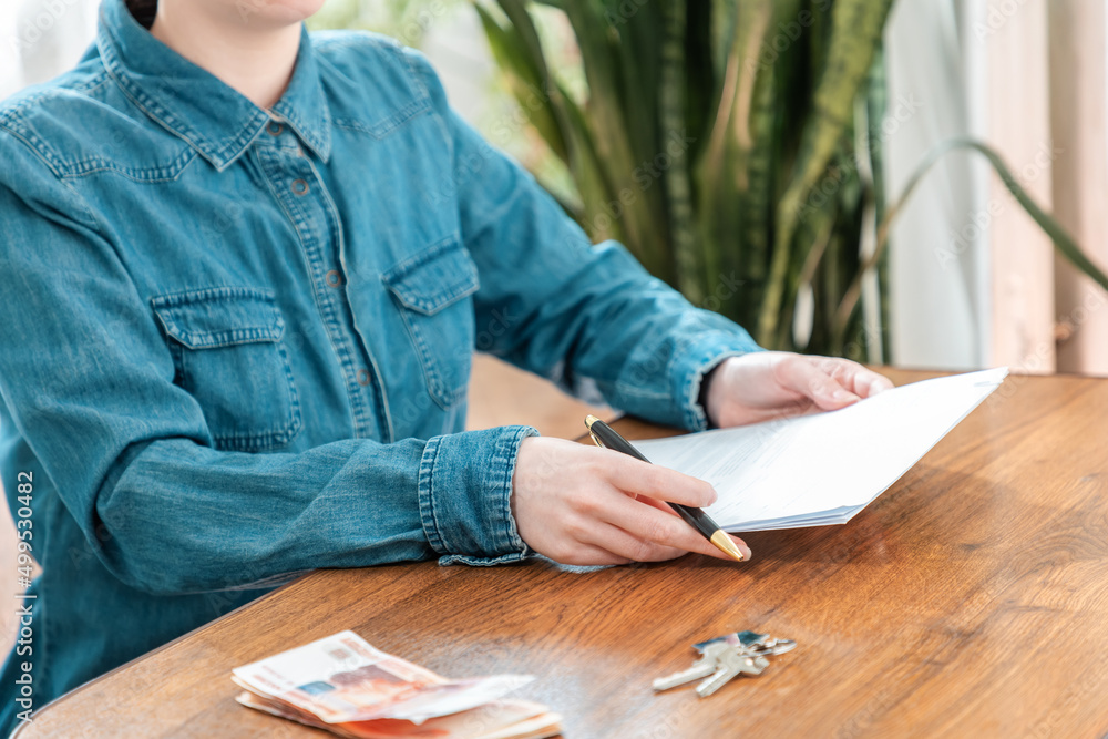 Close up of woman in a denim shirt holds documents and a pen in her hands. There is money and key on the table. The concept of insurance and mortgage Close up of woman in a denim shirt holds documents and a pen in her hands. There is money and key on the table. The concept of insurance and mortgage