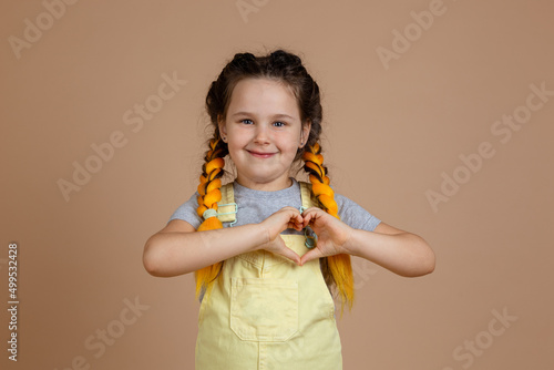 Little happy shining female child, having yellow kanekalon pigtails showing heart with fingers looking at camera wearing yellow jumpsuit and gray t-shirt on beige background.