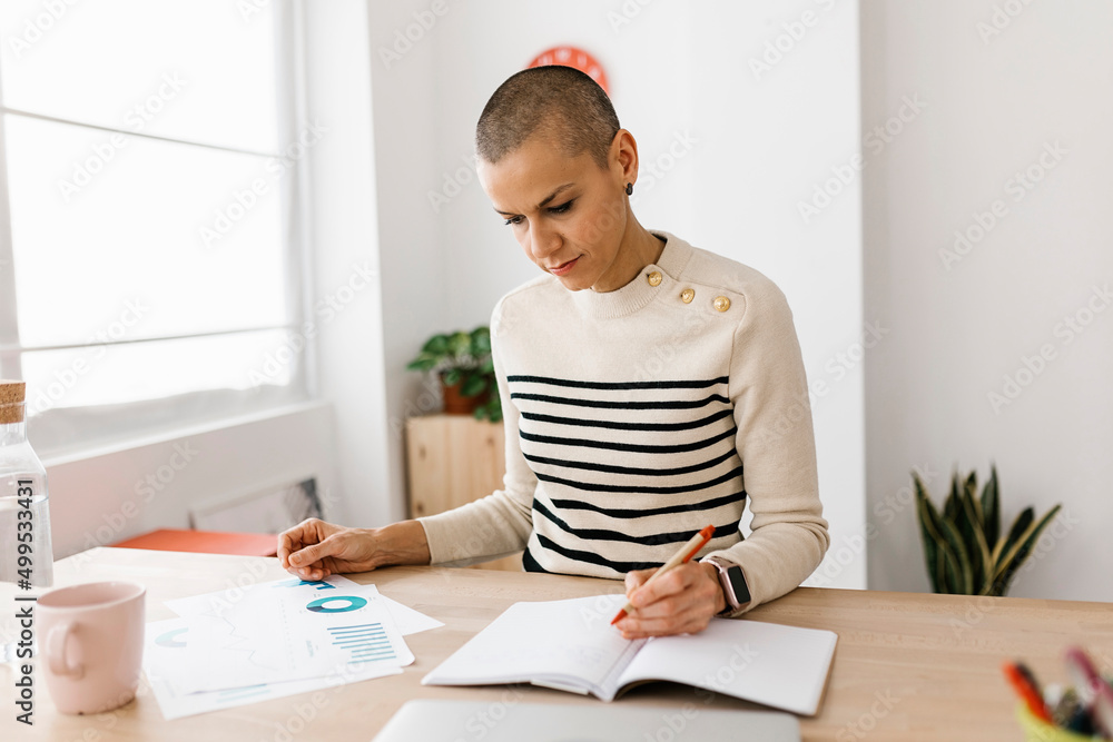 Mid adult woman analyzing and reviewing paperwork while working from home. Home office and business concept. Mid adult woman analyzing and reviewing paperwork while working from home. Home office and business concept.
