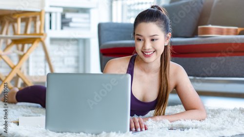 Millennial Asian young healthy slim strong sexy female sporty model in casual crop top sportswear laying down smiling on carpet floor in living room learning yoga workout class via laptop computer