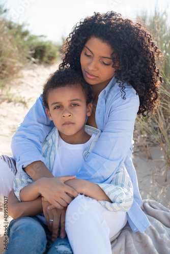 Tranquil African American mother and child on beach. Mother and son in casual clothes sitting on blanket, hugging. Family, relaxation, nature concept