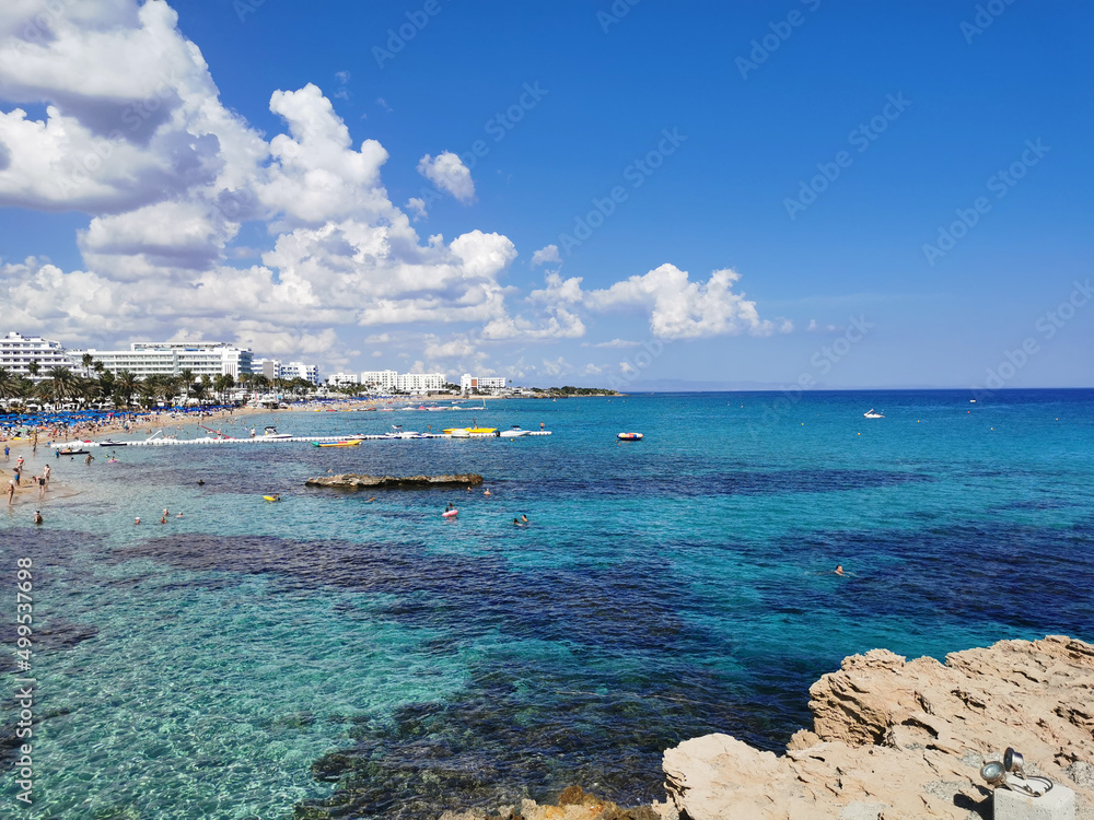 Large stones near the shore, clear water of the Mediterranean Sea, a view of the sandy large Sunrise beach and hotels against the background of a blue sky with clouds.