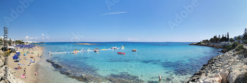 Protaras. Famagusta area. Cyprus. Panorama. Fig Tree Bay Beach, a small island opposite, people sunbathe and swim, catamarans and boats at the pier.