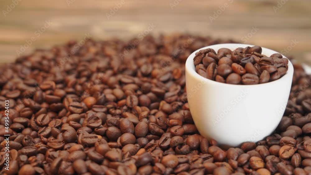 A white mug with coffee beans rotates standing against a background of roasted coffee beans. Front view, selective focus A white mug with coffee beans rotates standing against a background of roasted coffee beans. Front view, selective focus