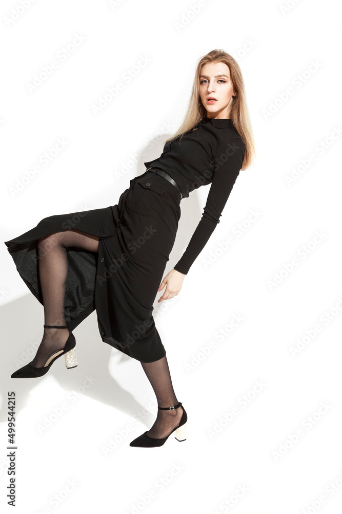 Full Length Portrait of Sensual Charming Female Blond Girl Posing in Alluring Long Black Dress With Lifted Leg Over White Background. Full Length Portrait of Sensual Charming Female Blond Girl Posing in Alluring Long Black Dress With Lifted Leg Over White Background.