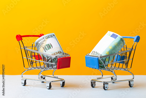 Savings Ideas. Two Shopping Trolleys Carts With US Dollars and Euro Currency Banknotes Over Trendy Yellow Background.