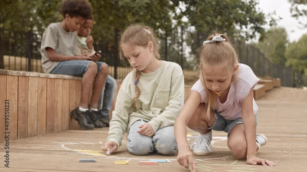 Slowmo of two pretty 10 year old Caucasian girls drawing with chalk on wooden planks while two African American boys playing games on smartphone in background, spending summertime outdoors in park Slowmo of two pretty 10 year old Caucasian girls drawing with chalk on wooden planks while two African American boys playing games on smartphone in background, spending summertime outdoors in park