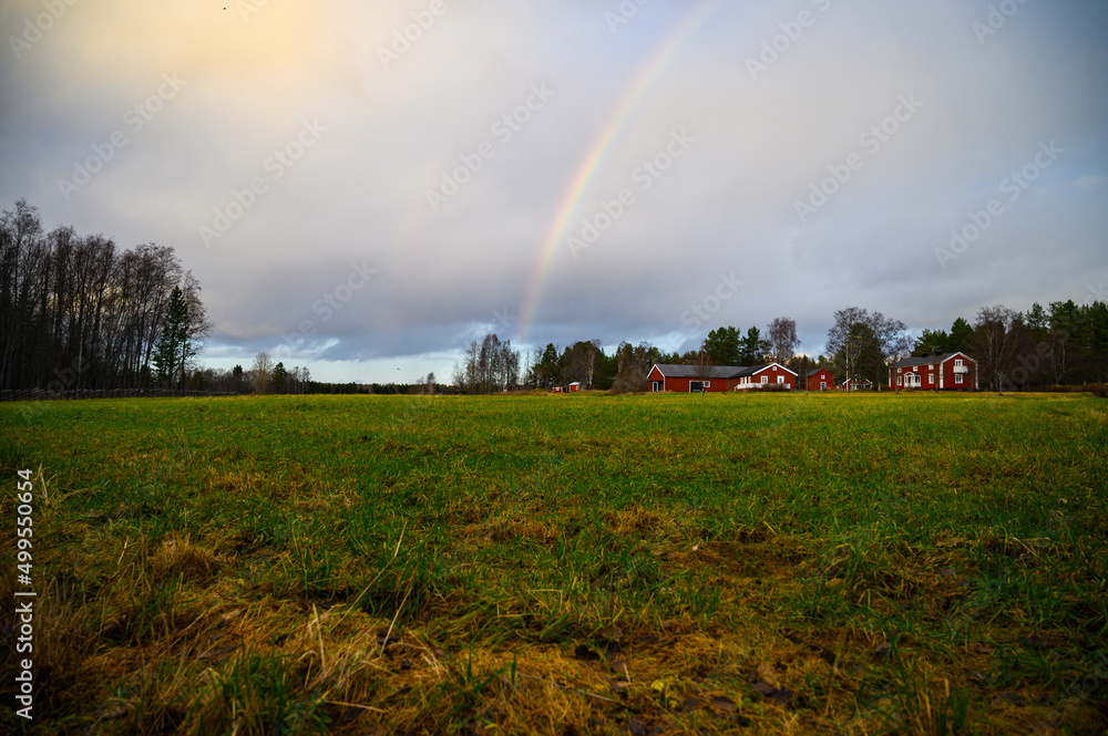 Naklejka premium A cloudy landscape view of open field filled with grass with cottages and rainbow in the sky in a rainy day during a outdoor visit