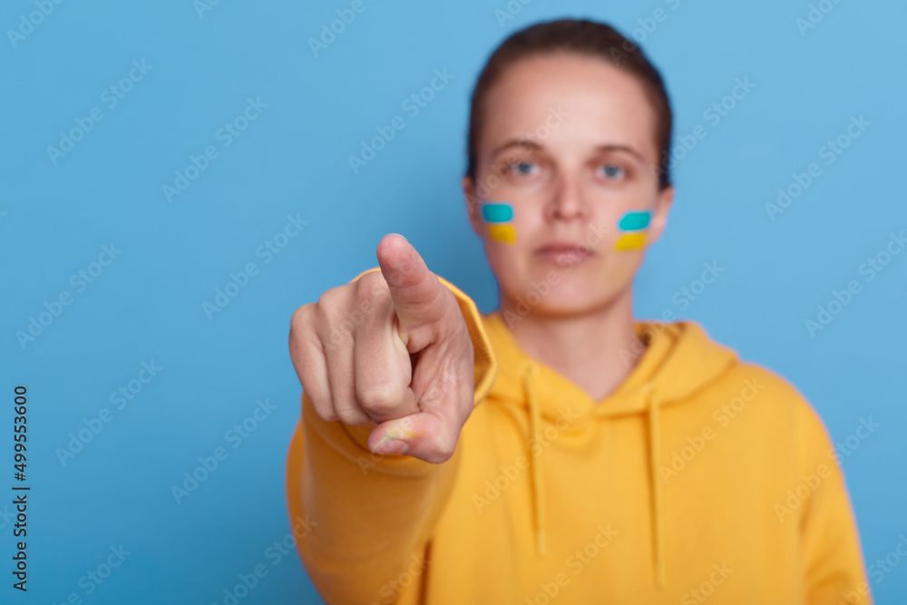 woman wearing yellow hoodie and with flag of Ukraine on her cheek, isolated over blue background, female pointing finger to camera, Stop fighting in Ukraine. Russia stop war.