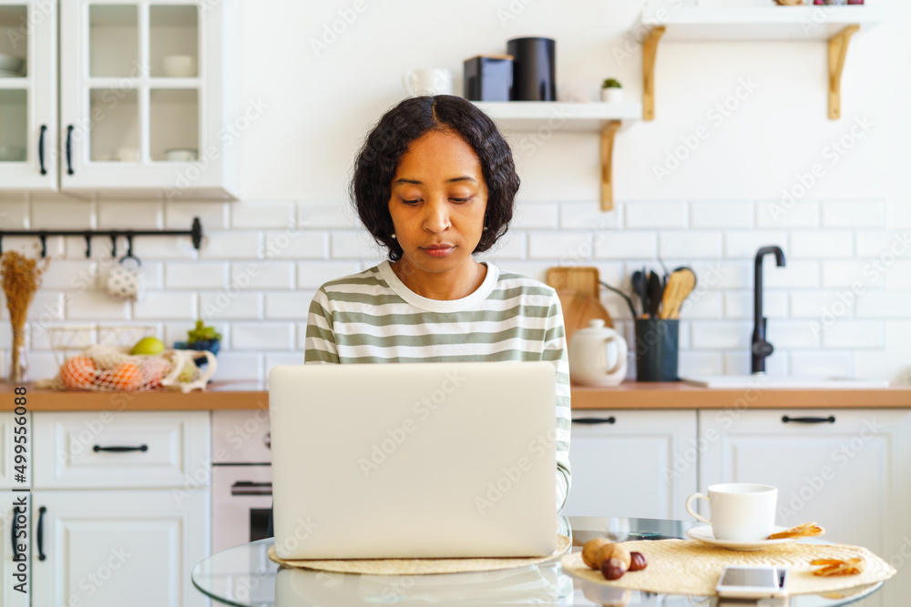Focused African-American female working from kitchen on laptop. Concept of home remote office Focused African-American female working from kitchen on laptop. Concept of home remote office