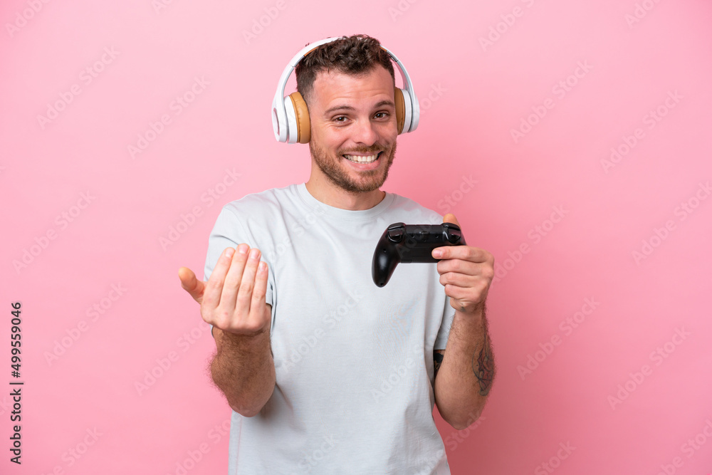Young Brazilian man playing with video game controller isolated on pink background inviting to come with hand. Happy that you came Young Brazilian man playing with video game controller isolated on pink background inviting to come with hand. Happy that you came