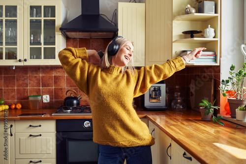 Joyful mid adult woman dancing in kitchen listening music on wireless headphones