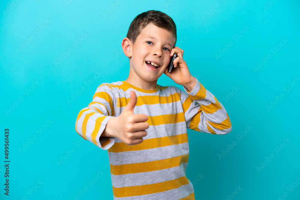 Little boy isolated on blue background keeping a conversation with the mobile while doing thumbs up Little boy isolated on blue background keeping a conversation with the mobile while doing thumbs up