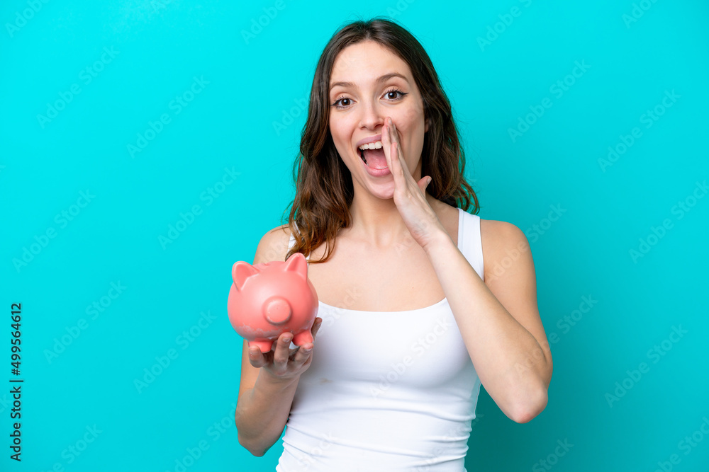 Young Caucasian woman holding a piggybank isolated on blue bakcground shouting with mouth wide open Young Caucasian woman holding a piggybank isolated on blue bakcground shouting with mouth wide open