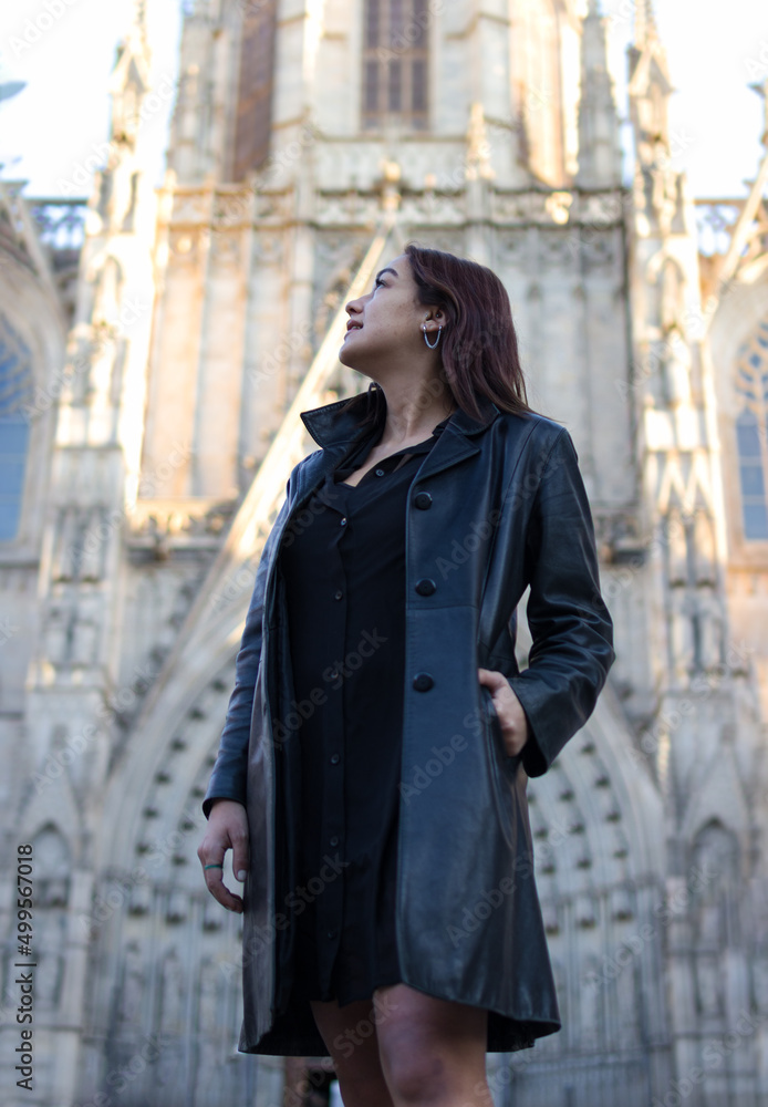 Fototapeta premium Three-quarter shot of a beautiful Latin woman looking up with one hand in a pocket somewhere in the city of Barcelona with an out of focus cathedral behind her