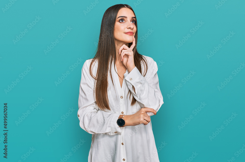 Young beautiful woman wearing casual white shirt with hand on chin thinking about question, pensive expression. smiling and thoughtful face. doubt concept. Young beautiful woman wearing casual white shirt with hand on chin thinking about question, pensive expression. smiling and thoughtful face. doubt concept.