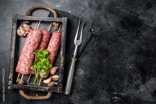 Fresh raw beef meat kebabs sausages on skewers in wooden tray. Black background. Top view. Copy space