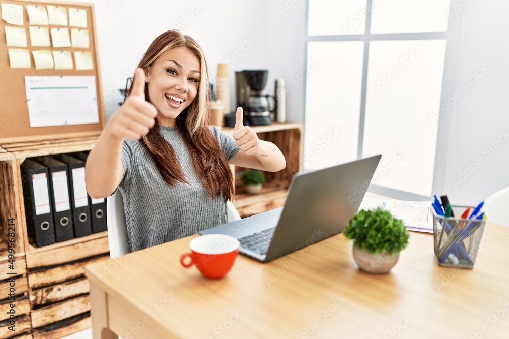 Young brunette woman working at the office with laptop approving doing positive gesture with hand, thumbs up smiling and happy for success. winner gesture. Young brunette woman working at the office with laptop approving doing positive gesture with hand, thumbs up smiling and happy for success. winner gesture.