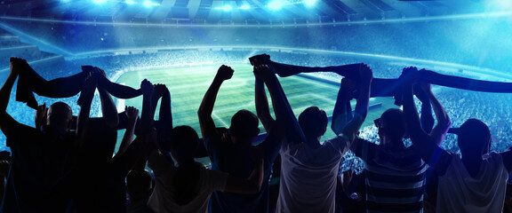 Back view of football, soccer fans cheering their team with state flags and scarfs at crowded stadium at evening time. Concept of sport, support, competition. Out of focus effect