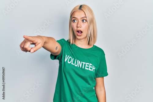 Beautiful blonde woman wearing volunteer t shirt pointing with finger surprised ahead, open mouth amazed expression, something on the front