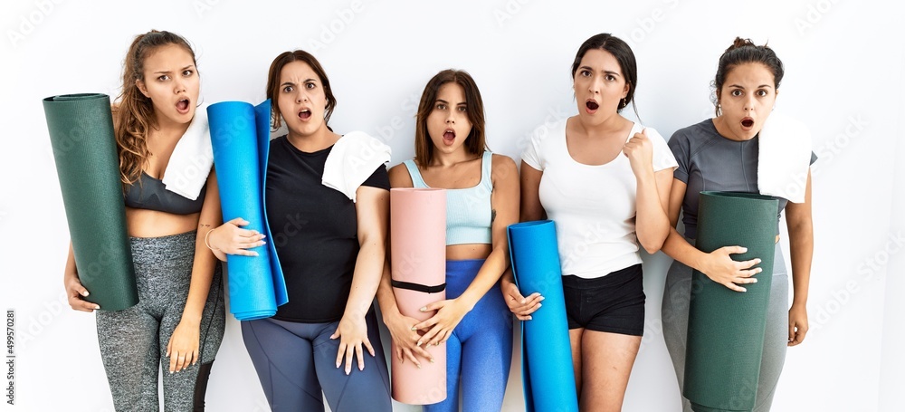 Group of women holding yoga mat standing over isolated background in shock face, looking skeptical and sarcastic, surprised with open mouth