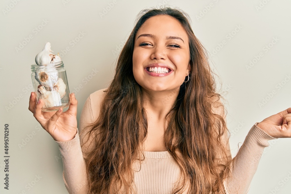 Young hispanic girl holding glass of garlic screaming proud, celebrating victory and success very excited with raised arm Young hispanic girl holding glass of garlic screaming proud, celebrating victory and success very excited with raised arm