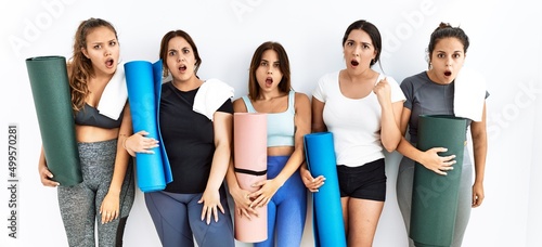 Group of women holding yoga mat standing over isolated background in shock face, looking skeptical and sarcastic, surprised with open mouth