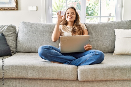 Photography Beautiful young brunette woman sitting on the sofa using computer laptop at home smiling positive doing ok sign with hand and fingers